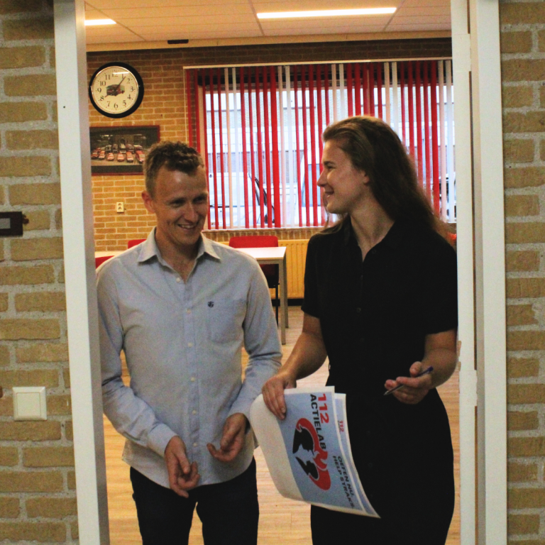 Twee mensen lopen door een deur, een vrouw houdt een document vast en lacht naar een man.